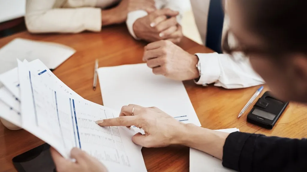 Two professionals reviewing printed documents and charts at a wooden table.