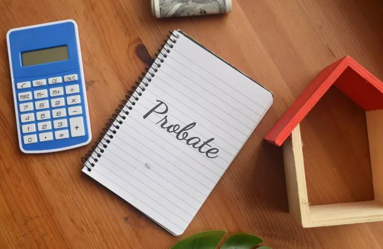 Spiral notebook with "Probate" text, blue calculator, rolled dollar bill, and small wooden house model on wooden table.