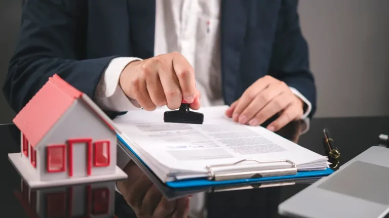 Person in business attire stamping documents on a desk with a small red and white house model nearby.