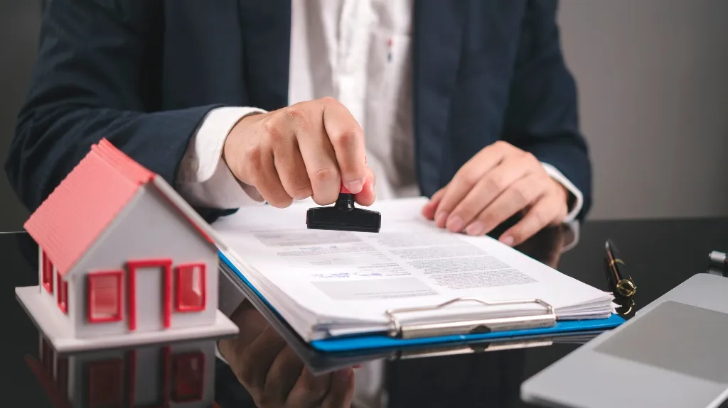 Person in business attire stamping documents on a desk with a small red and white house model nearby.