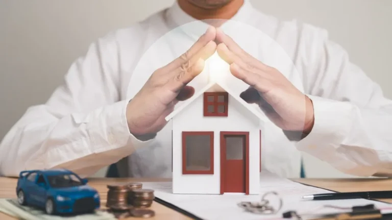 Person in white shirt protecting small house model with hands on desk with coins, keys, and toy car.