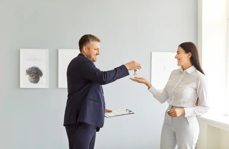 Man in dark suit handing over keys to smiling woman in white blouse in bright room.