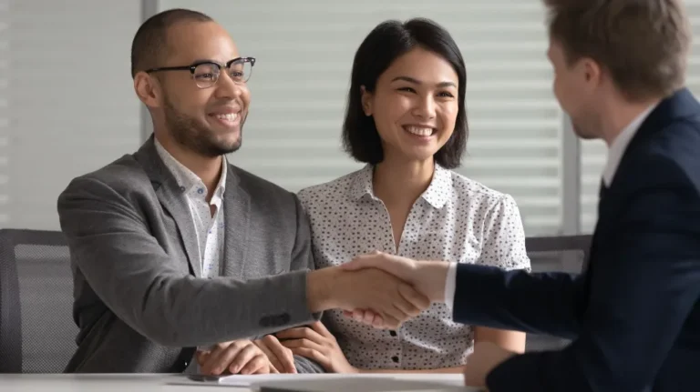 Three professionally dressed individuals, two men and one woman, shaking hands and smiling in an office setting.