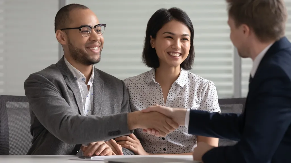 Three professionally dressed individuals, two men and one woman, shaking hands and smiling in an office setting.