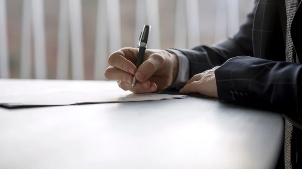 Close-up of a person in a dark suit writing on paper with a black pen at a desk.