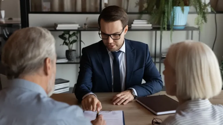 Businessman in navy suit and glasses explaining document to senior couple at office desk.