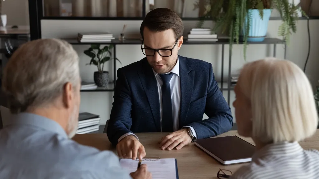 Businessman in navy suit and glasses explaining document to senior couple at office desk.