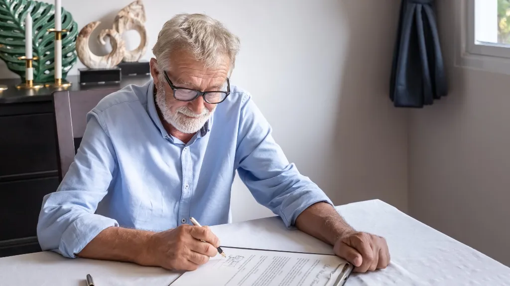 Elderly man with glasses signing a document at a white table indoors.