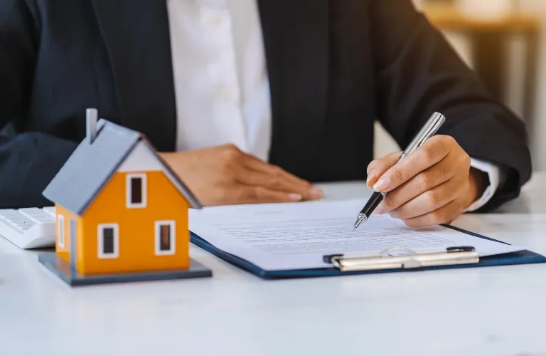 Person in business attire signing a document on clipboard with a small orange house model nearby.