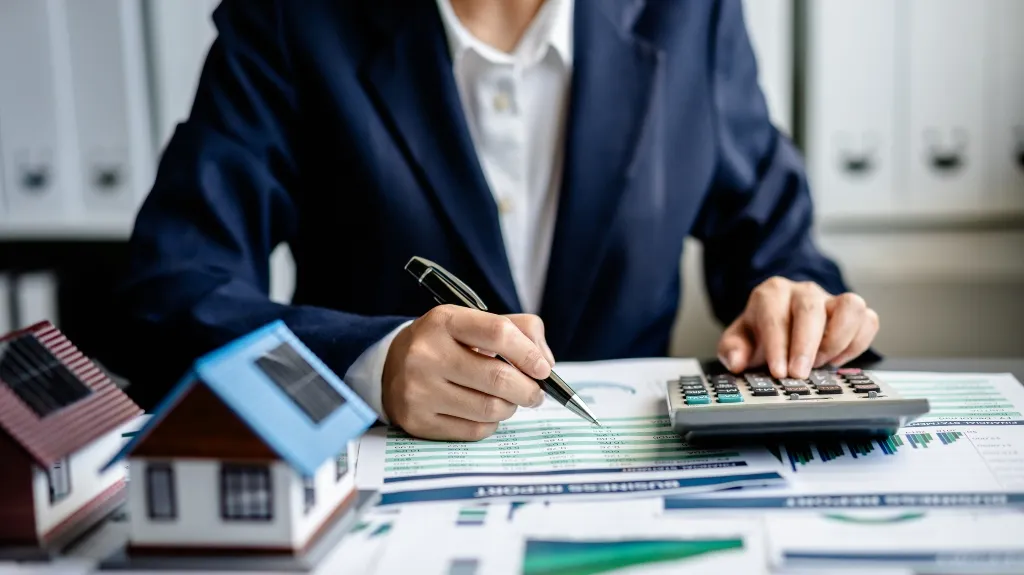 Person in dark suit using calculator and pen, analyzing financial documents with house models.