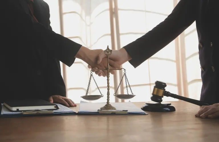 Two individuals in dark suits shaking hands over a wooden table with a balance scale and judge's gavel.